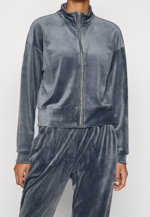 Osoba ubrana w szarą aksamitną kurtkę na zamek błyskawiczny oraz pasujące spodnie z długimi rękawami i bocznymi kieszeniami, stojąca na jednolitym tle.