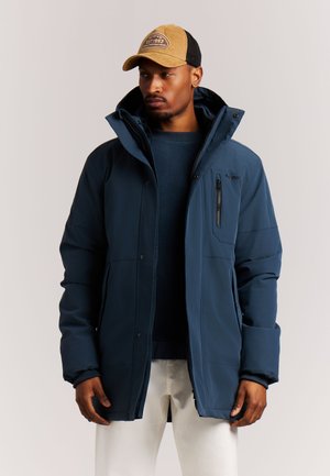 Man die een blauwe winterjas met hoge kraag, marineblauwe trui, witte broek en een beige-zwarte baseballpet draagt en opzij kijkt.