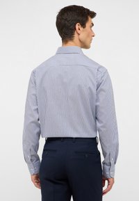 Vista posteriore di una camicia a maniche lunghe con sottili righe blu e bianche, vestibilità slim, polsini con bottoni e colletto classico. Pantaloni blu navy.