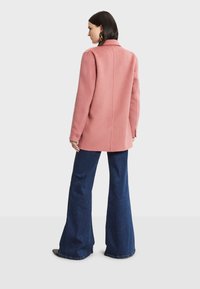 Abrigo rosa de mezcla de lana con corte recto, cuello con muescas y mangas largas, combinado con pantalones de mezclilla acampanados. Textura suave y diseño sencillo.