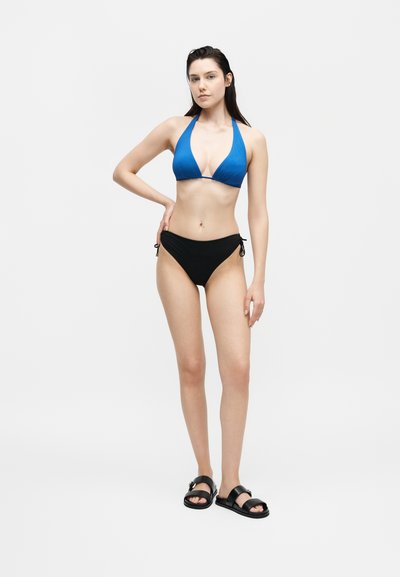 Νέα γυναίκα που φορά μπλε μπικίνι τοπ με λαιμόκοψη halter, μαύρο κάτω μέρος μπικίνι και μαύρα σανδάλια σε λευκό φόντο.