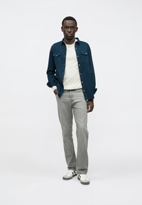 Neformálny outfit zahŕňajúci tmavomodrú košeľu, krémový sveter, sivé džínsy a bielo-čierne tenisky, prezentujúci moderný vrstvený dizajn.