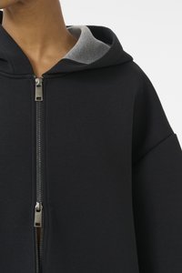 Zwarte zip-up sweatshirt met een grijze capuchonvoering, met een grote, relaxte pasvorm en een gladde textuur. Ritsdetail zichtbaar aan de voorzijde.
