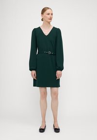 Zelena haljina s V-izrezom, dugim napuštenim rukavima i pojasom. Glatka tkanina, dužine do koljena, minimalistički dizajn i bez vidljivih uzoraka.