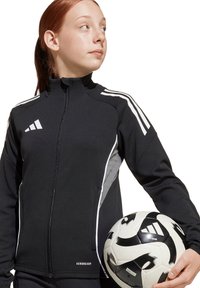 Zwarte sportjack met witte accenten, voorzipper, hoge kraag en details op de zijpanelen, gecombineerd met een zwart-wit voetbal.