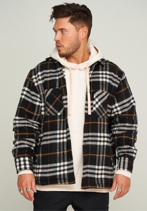Schwarz-weiße Karojacke mit orangefarbenen Akzenten, die zwei Brusttaschen und eine weiche Textur aufweist, über einem hellen Hoodie getragen.