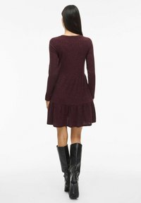 Robes maroon à côtes avec des manches longues et un ourlet évasé. Portée avec des bottes noires à talons montant jusqu'aux genoux, mettant en valeur une taille cintrée et une jupe superposée.