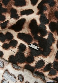 Gros plan sur un tissu imprimé léopard avec une petite étiquette métallique rectangulaire gravée du mot « NEXT » fixée près d'une couture cousue.