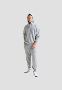 Felpa grigia con cappuccio, chiusura con cordino e polsini a coste abbinata a pantaloni della tuta coordinati. Ai piedi ci sono sneakers bianche con dettagli blu navy.