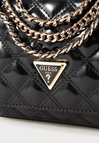 Black quilted handbag with a glossy finish, featuring dual gold-tone chain straps and a triangular logo plaque with "GUESS U.S.A." text.