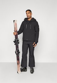 Schwarze isolierte Skijacke mit Reißverschluss-Taschen, kombiniert mit passenden Skihosen. Das Modell hält Skier mit einem Farbverlauf-Design und Bindungen.