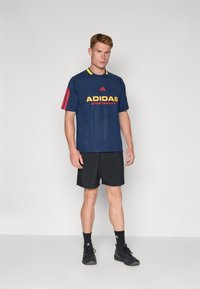 Jūrinės sportinės marškinėliai su raudonais ir geltonais akcentais, kuriuose matomas "ADIDAS SPORTSWEAR" logotipas. Dėvimi su juodomis šortais ir juodais sportiniais batais.