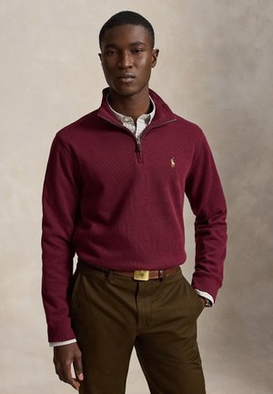 Homme portant un pull bordeaux à fermeture éclair partielle sur une chemise à carreaux, avec un pantalon marron et une ceinture, debout devant un fond uni.