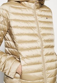 Pletená bunda v metalické zlaté barvě, se vodorovným prošíváním, zipem vpředu a kapucí. Obsahuje boční kapsy a akcent s logem.