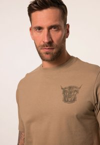Beige t-shirt met korte mouwen van zachte katoen, met een grafische print op de linker borst die een gestileerd schildontwerp weergeeft.