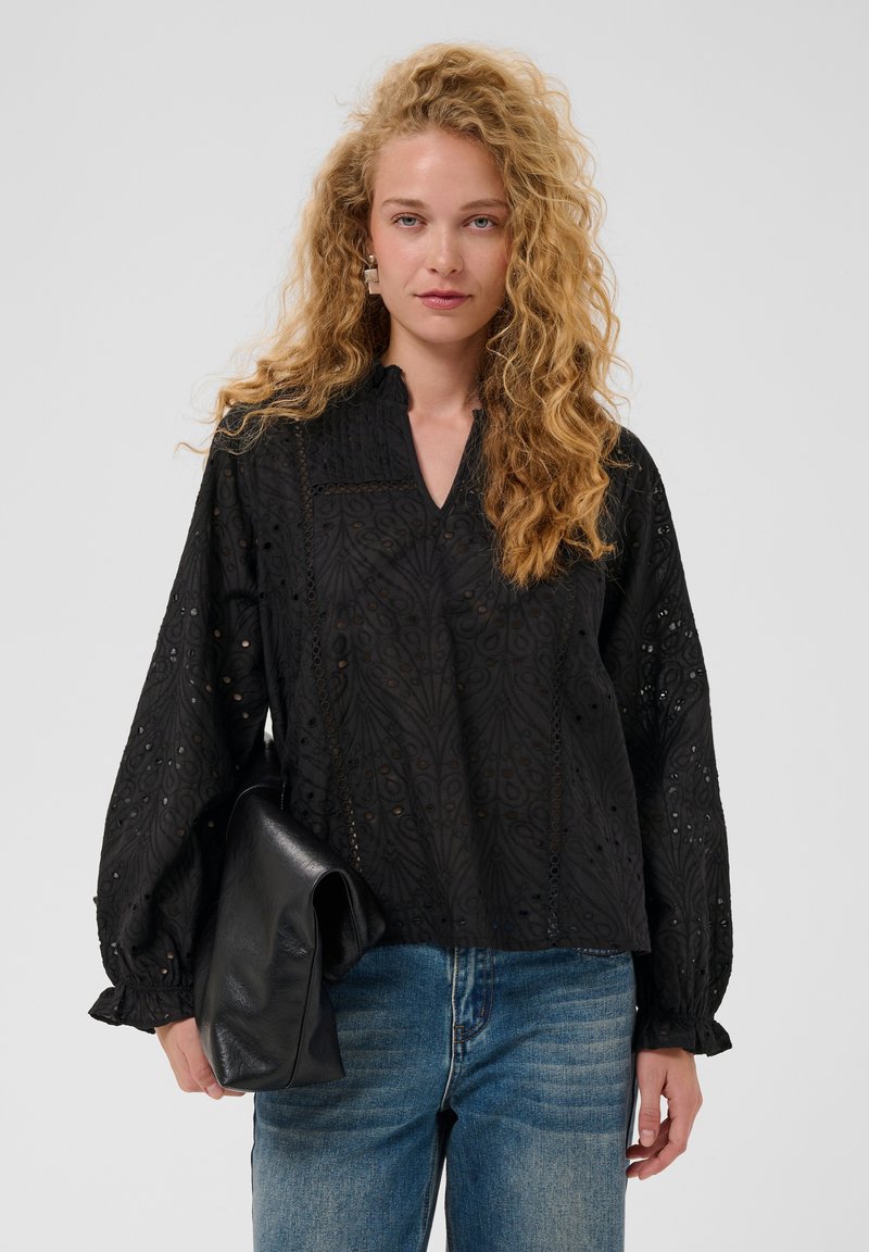 Femme aux longs cheveux blonds bouclés, portant un chemisier noir à motifs et un jean bleu, tenant une pochette en cuir noire pliée.