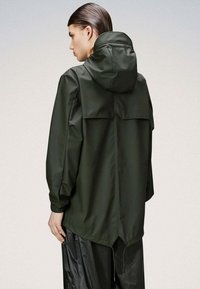 Veste imperméable verte avec capuche, présentant un empiècement au dos et des poignets élastiques. Texture lisse et détails de ourlet réglables.