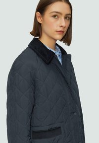 Veste noire matelassée avec un col en velours côtelé, comportant des boutons sur le devant et des poches latérales, présentée sur un fond clair.