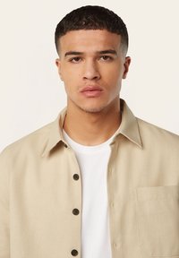 Beige overhemd met knopen en een kraag, voorzien van een voorzak en donkere knopen, gedragen over een wit T-shirt. Soepele, gestructureerde stof.