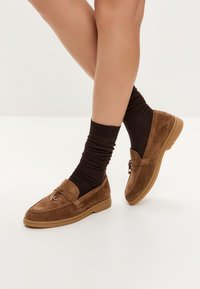 Mocasines de ante marrón con suela de goma, que cuentan con un acento decorativo y se combinan con calcetines de canalé oscuros. El diseño es bajo y casual.