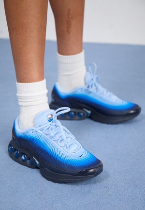 Nogi w białych skarpetkach i niebieskich sneakersach Nike Air Max z widoczną poduszką powietrzną na jasnoniebieskiej podłodze.