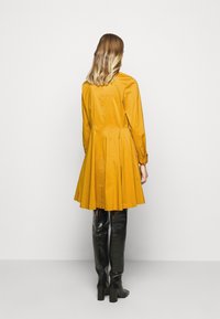 Sukienka w musztardowym kolorze z dopasowanym gorsetem, rozszerzanym dołem, długimi rękawami i detalami w postaci guzików, zestawiona z czarnymi skórzanymi botkami do kolan.