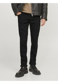 Svarta jeans i smal passform med en slät textur, kombinerade med en brun tröja och en svart läderjacka. Skorna är svarta snörkängor.