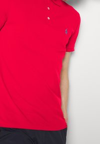 Červené polo tričko z bavlny se zapínáním na tři knoflíky, krátkými rukávy a modrým logem na levé straně hrudi. Hladká textura a přiléhavý střih.