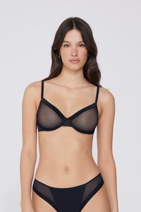 Femme aux longs cheveux foncés portant un soutien-gorge noir transparent et une culotte noire assortie, debout devant un fond clair uni.