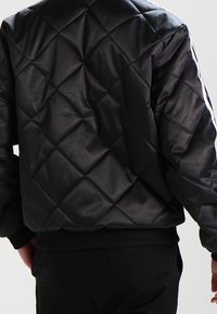 Chaqueta bomber acolchada negra, confeccionada en material brillante, con ribetes en el bajo y los puños de las mangas, y rayas blancas a lo largo de los hombros.
