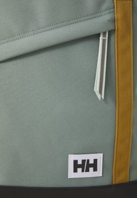 Zaino grigio chiaro con superficie texture, dotato di spallacci gialli, zip superiore e una patch con logo bianco nella parte inferiore.