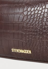 Hnedý kožený povrch s textúrou krokodílej kože a zlatým detailom loga "Steve Madden" pri dolnom okraji.
