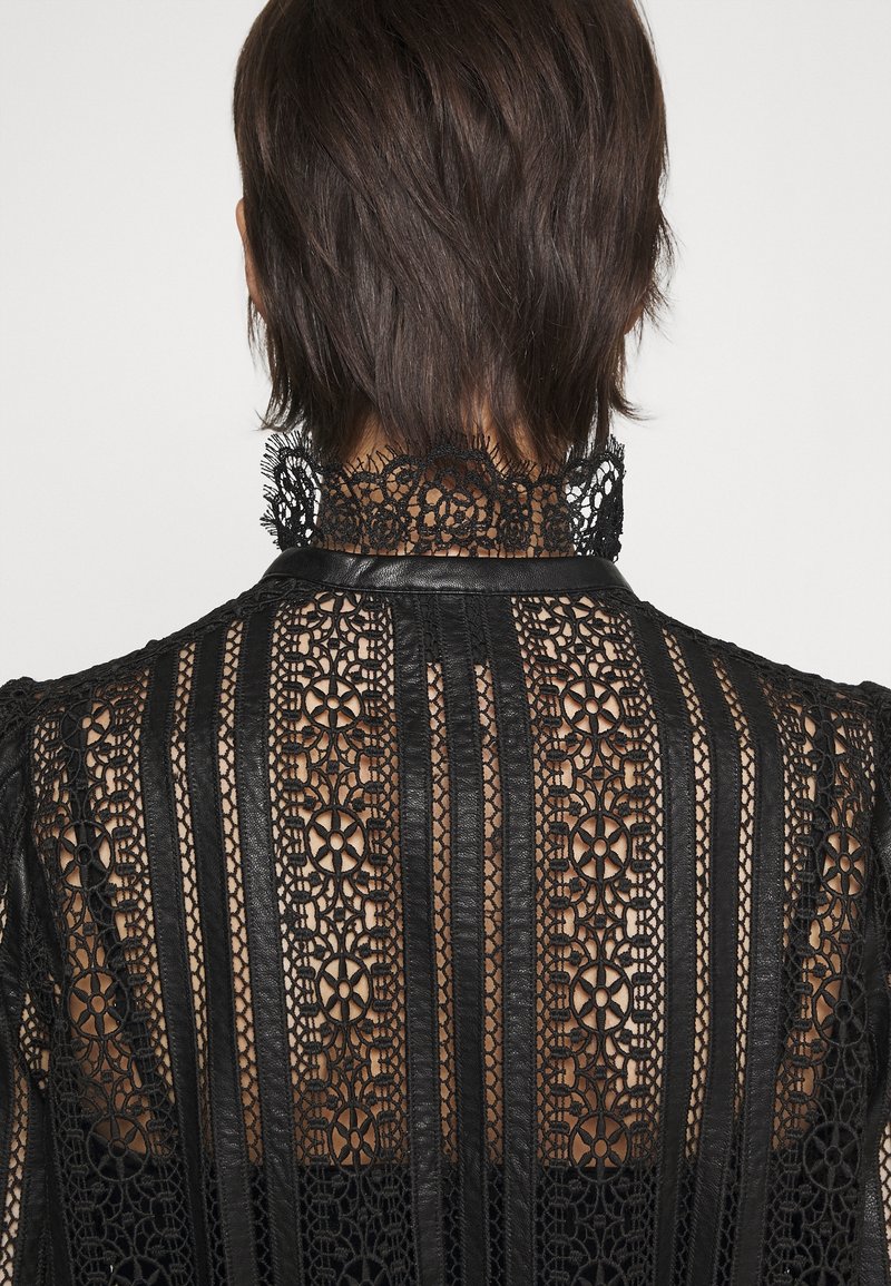 Haut en dentelle noire présentant des motifs floraux complexes avec un col montant, des accents en cuir et des rayures verticales, révélant un design texturé.