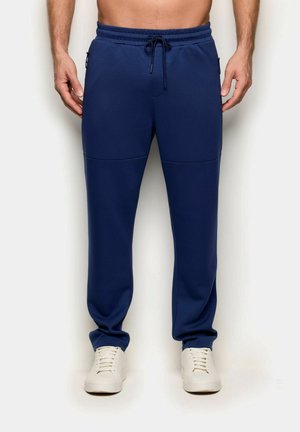 Uomo che indossa pantaloni da jogging blu navy con vita con coulisse e sneakers bianche, in piedi contro uno sfondo chiaro e semplice.