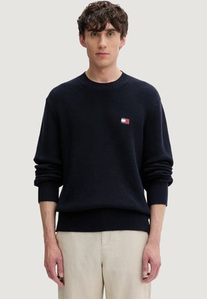 Jeune homme aux cheveux bouclés et foncés, portant un pull en maille noir et un pantalon beige clair, debout devant un fond clair uni.