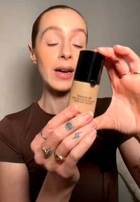 Person with closed eyes holds beige foundation bottle with black cap, showing it close to the camera, wearing brown shirt and multiple rings.