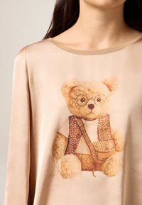 Top beige a maniche lunghe con grafica di un orso. L'orso indossa occhiali, un gilet con borchie e ha un aspetto morbido e strutturato.