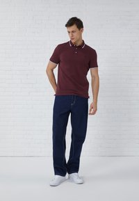 Burgunderfarbenes Polohemd mit weißem Besatz, kurzen Ärmeln und einem Logo auf der Brust, kombiniert mit dunkelblauen Jeans und weißen Sneakers.