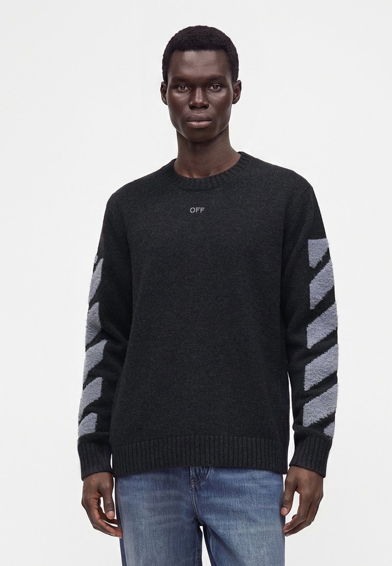 Homme adulte portant un pull noir avec des rayures diagonales grises sur les manches et un petit texte "OFF" sur la poitrine, associé à un jean bleu, debout.