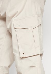 Beige cargo kalhoty vyrobené z lehkého materiálu. Mají velkou boční kapsu s klopou, viditelné švy a kovový patent.