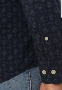 Mörkblå skjorta med en texturerad tyg som har ett upprepat mönster av diamanter och virvlar. Knappdetalj på ärmen, med en rak nederkant.