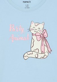 Gato creme sorridente com manchas cor-de-rosa e uma grande laço rosa brilhante, ao lado de texto brilhante em rosa com a frase "Animal de Festa" sobre um tecido azul claro.