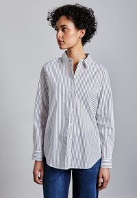 Kiharatukkainen nainen valkoisessa, mustaraidallisessa nappipaidassa ja sinisissä farkuissa, katsoen vasemmalle yksiväristä taustaa vasten.