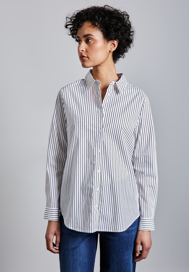 Kiharatukkainen nainen valkoisessa, mustaraidallisessa nappipaidassa ja sinisissä farkuissa, katsoen vasemmalle yksiväristä taustaa vasten.