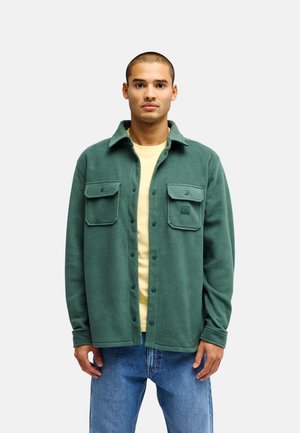Junger Mann mit grüner Fleece-Hemdenjacke, gelbem Hemd und blauen Jeans, stehend vor einem schlichten hellen Hintergrund.