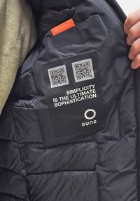Interno di una giacca nera imbottita con un'etichetta in tessuto contenente codici QR. È visibile un'etichetta rossa con la dicitura "SAFE POCKET" vicino a un'area in pile color beige.
