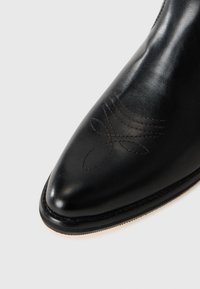 Botín negro de cuero hasta el tobillo con punta afilada y un patrón decorativo de costura marrón en el lateral. Textura suave con finos detalles a lo largo del borde.