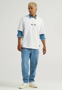 Biała, oversize'owa koszulka z denimowym kołnierzem, z czarnym logo "Lee". W połączeniu z niebieskimi spodniami w paski i białymi sneakersami.