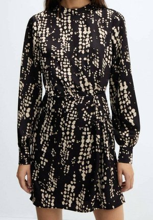 Robe noire à manches longues avec des taches abstraites beiges, col montant, taille cintrée et manches à poignets, portée par une personne debout.