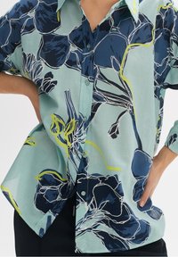 Light blue collared shirt featuring a floral pattern in navy and yellow accents; button-up front; loose fit with rolled-up sleeves.